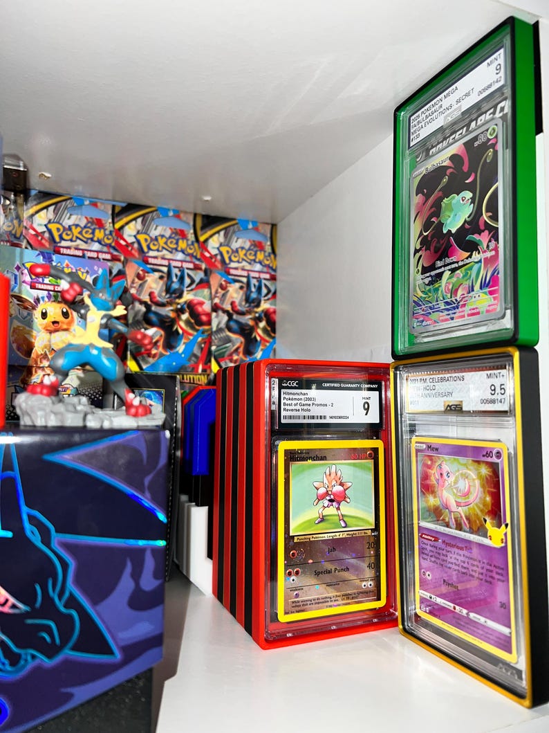 May include: A display of graded Pok&eacute;mon trading cards in protective cases. The cases are various colors, including green, red, and black. The cards feature images of Pok&eacute;mon characters like Hitmonchan and Mew. Background includes Pok&eacute;mon booster packs and a Lucario figurine.