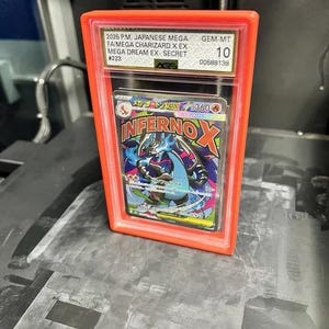 May include: A graded Japanese Mega Charizard X EX card in a red protective case. The card features a colorful illustration of a dragon-like creature with the text "Inferno X". The case is labeled with details including "GEM-MT 10".