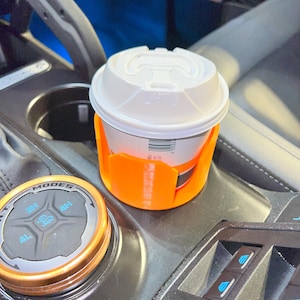 May include: An orange cup holder designed for a vehicle's center console, holding a white coffee cup with a lid. The cup holder is made of plastic and is designed to fit snugly in the vehicle's cup holder slot.