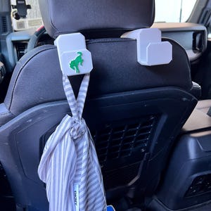 May include: White car seat headrest hooks with a green horse logo and a gray and white striped bag. The bag is knotted and has the DKNY logo. The hooks are attached to the car seat.