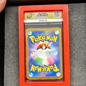 May include: A graded Pok&eacute;mon trading card encased in a clear plastic holder, set within a red frame. The card features the Pok&eacute;mon logo and a colorful design. The holder is labeled with "AGS" and a serial number.