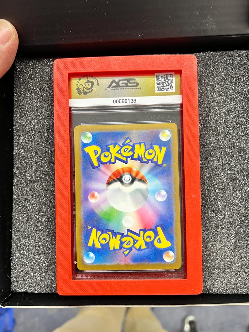 May include: A graded Pok&eacute;mon trading card encased in a clear plastic holder, set within a red frame. The card features the Pok&eacute;mon logo and a colorful design. The holder is labeled with "AGS" and a serial number.