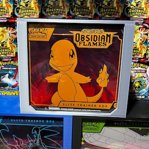 May include: An Elite Trainer Box featuring the Pokémon character Charmander in orange, with the text "Obsidian Flames" and "Trading Card Game." The box has a silver frame and is surrounded by other Pokémon card packs.