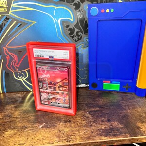 May include: A red graded card holder containing a Pokemon card with a colorful illustration of a town. A blue electronic device and a Pokemon box are in the background. The card holder is on a wooden surface.
