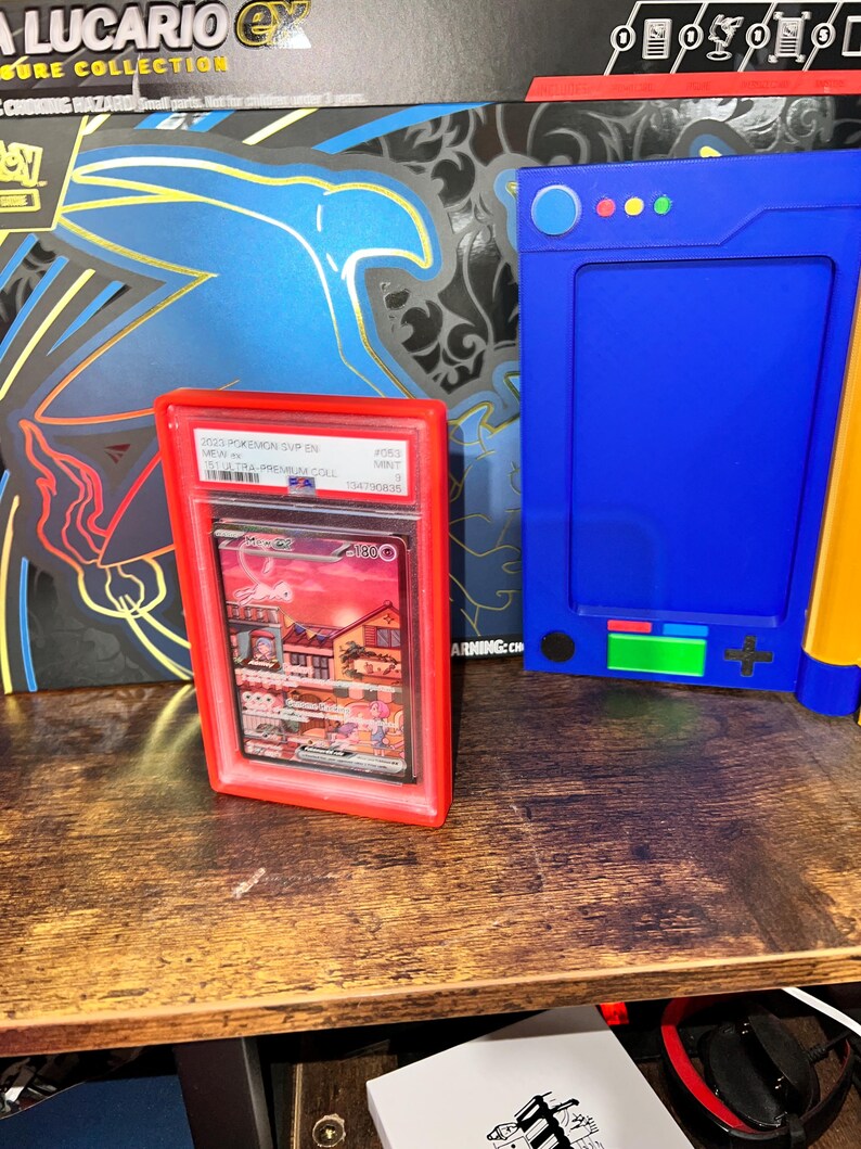 May include: A red graded card holder containing a Pokemon card with a colorful illustration of a town. A blue electronic device and a Pokemon box are in the background. The card holder is on a wooden surface.