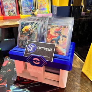May include: A blue and white card storage box with the text "SantiWorks" and a logo. The box holds several trading cards in protective cases. Other graded cards are displayed in the background.