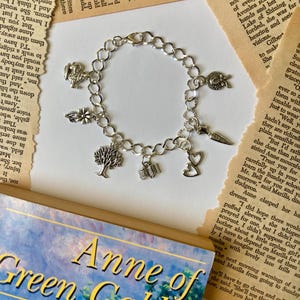 May include: Silver charm bracelet with various charms, including a tree, book, and heart. The bracelet is displayed with a copy of "Anne of Green Gables" and vintage book pages.