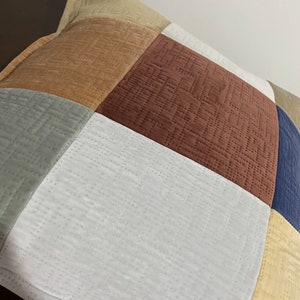 Pode incluir: Uma almofada quadrada com um design de patchwork. A almofada é feita de tecidos de diferentes cores, incluindo bege, marrom, cinza, branco e azul.