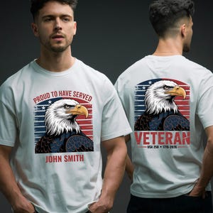 Op de afbeelding: Wit T-shirt met een gedetailleerde illustratie van een Amerikaanse zeearend tegen een rood, wit en blauwe achtergrond. De voorkant toont "PROUD TO HAVE SERVED" en de naam "JOHN SMITH". De achterkant zegt "VETERAN".