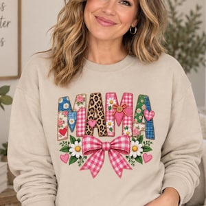 Könnte beinhalten: Ein beigefarbenes Sweatshirt mit Rundhalsausschnitt und dem Wort MAMA in großen, gemusterten Buchstaben, darunter Blumenmuster, Leopardenmuster und rosa Vichy-Karo. Eine große rosa Vichy-Schleife, weiße Gänseblümchen und kleine rosa Herzen zieren den unteren Bereich.