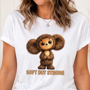 Puede incluir: Una camiseta blanca de manga corta con el gráfico de una criatura marrón, peluda y de orejas grandes con ojos grandes. Debajo de la criatura, el texto "SOFT BUT STRONG" está impreso en una fuente naranja claro con borde oscuro.