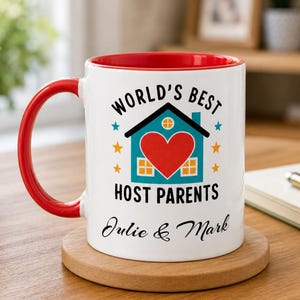 Pode incluir: Caneca de cerâmica branca com interior e alça vermelhos. A caneca apresenta o texto "WORLD'S BEST HOST PARENTS" com um gráfico de casa e os nomes "Julie & Mark". A caneca está sobre um descanso de copo de madeira.