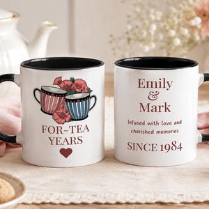 May include: Two white ceramic mugs with black rims and handles. One mug features a graphic of two teacups, red flowers, a heart, and the text FOR-TEA YEARS. The other mug displays the text Emily & Mark, Infused with love and cherished memories, and SINCE 1984.