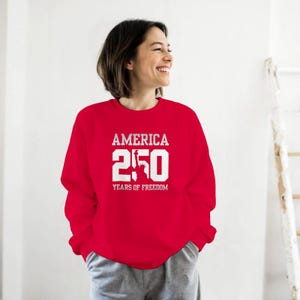 Puede incluir: Una sudadera roja con el texto blanco "AMERICA 250 YEARS OF FREEDOM". El dise&ntilde;o incluye el n&uacute;mero 250 y una imagen de la Estatua de la Libertad. La sudadera es de cuello redondo.