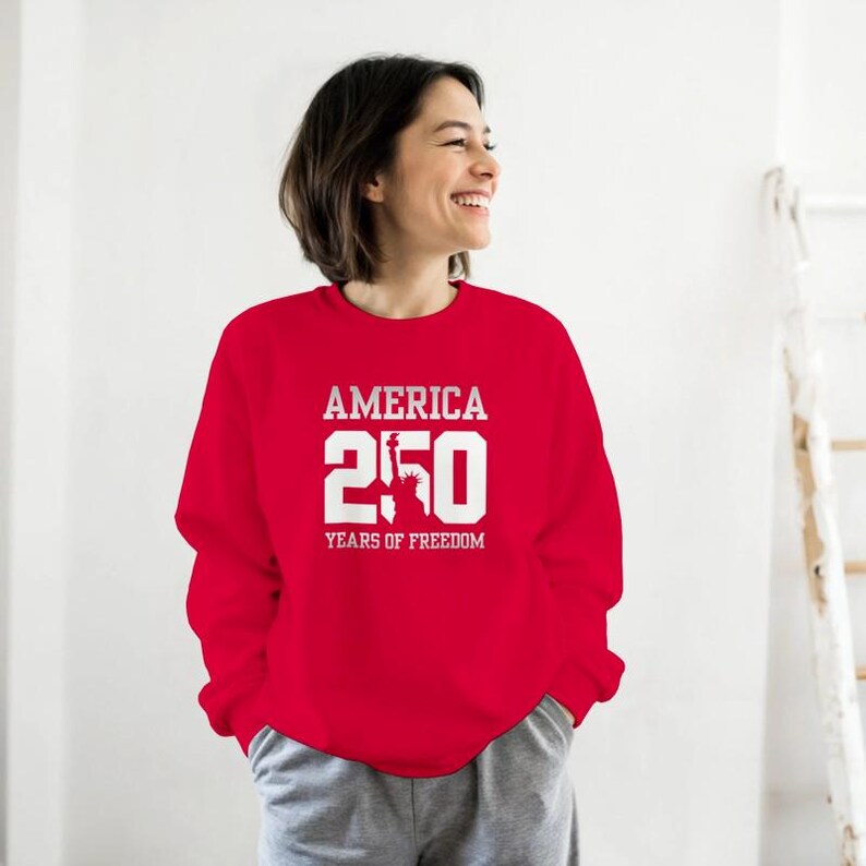 Puede incluir: Una sudadera roja con el texto blanco "AMERICA 250 YEARS OF FREEDOM". El dise&ntilde;o incluye el n&uacute;mero 250 y una imagen de la Estatua de la Libertad. La sudadera es de cuello redondo.
