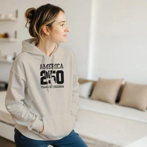 Puede incluir: Una sudadera con capucha color crema con un bolsillo delantero y una capucha con cord&oacute;n. La parte delantera de la sudadera con capucha presenta el texto "AMERICA 250 YEARS OF FREEDOM" en negrita, en negro. La persona de la imagen lleva la sudadera.
