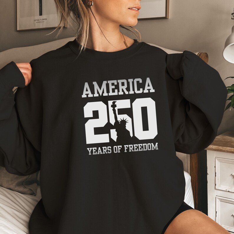 Puede incluir: Sudadera negra con el texto blanco "AMERICA 250 YEARS OF FREEDOM". El dise&ntilde;o incluye el n&uacute;mero "250" y la silueta de la Estatua de la Libertad. Prenda de tem&aacute;tica patri&oacute;tica.