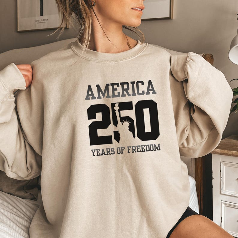 Puede incluir: Sudadera beige con el texto "AMERICA 250 YEARS OF FREEDOM" en negro y gris. El n&uacute;mero "250" presenta la silueta de la Estatua de la Libertad. La sudadera es de cuello redondo.