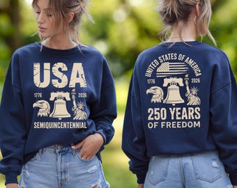May include: A navy blue long-sleeve sweatshirt featuring a cream-colored graphic. The front displays USA, 1776-2026, Semiquincentennial, an eagle, the Liberty Bell, and the Statue of Liberty. The back shows United States of America, 250 Years of Freedom.