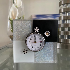 May include: A white and black clock with a silver face and black hands. The clock is on a white and black glittery base with white flowers.