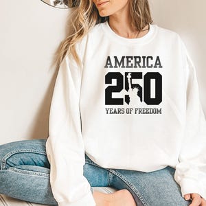 Puede incluir: Sudadera blanca con el texto "AMERICA 250 YEARS OF FREEDOM" en negro. El n&uacute;mero 250 incluye una silueta de la Estatua de la Libertad. La sudadera es de estilo casual.