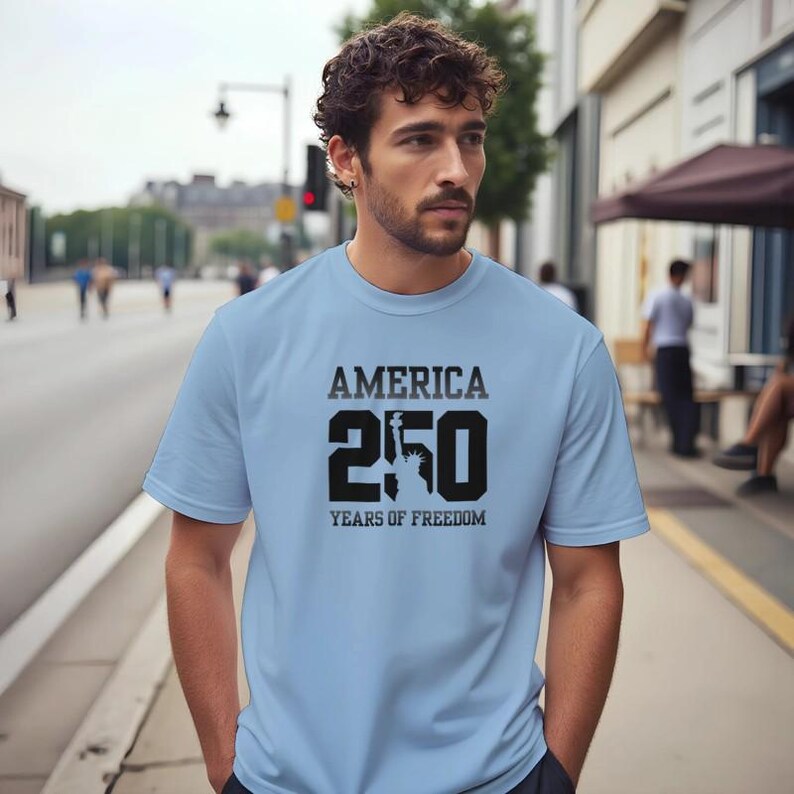Puede incluir: Camiseta azul claro con el texto "AMERICA 250 YEARS OF FREEDOM" en negro. El n&uacute;mero 250 incluye una silueta de la Estatua de la Libertad. La camiseta es de material suave.
