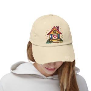 Peut inclure: Casquette de baseball beige délavé avec un motif de maison colorée et un cœur dans la fenêtre. Le texte "World's Best Host Mum" est en dessous. La casquette a un aspect usé.