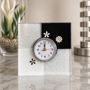 May include: A square quartz clock with a black, white, and glittery silver design. The clock face is white with black numbers and hands. Small white flower accents are on the clock's face. The clock is on a marble surface.