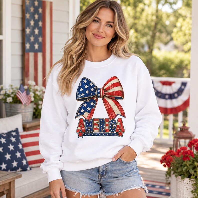 May include: A white long-sleeve sweatshirt featuring a large graphic of a bow with a red, white, and blue star and stripe pattern. Below the bow, the word AMERICA is printed in bold, block letters with a matching patriotic color scheme.