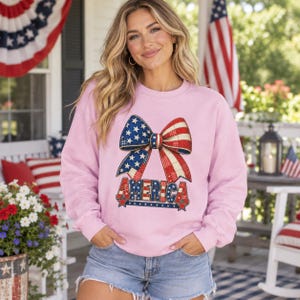May include: A light pink long-sleeve sweatshirt featuring a large graphic of a bow with a red, white, and blue star and stripe pattern. The word AMERICA is printed in a matching patriotic font below the bow, also decorated with stars and stripes.