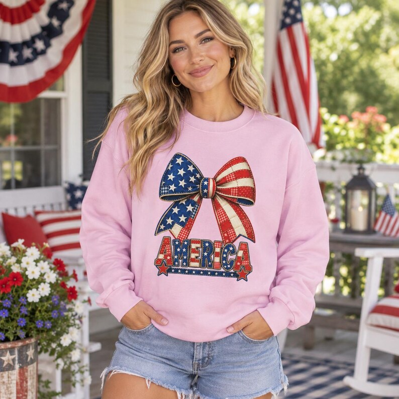 May include: A light pink long-sleeve sweatshirt featuring a large graphic of a bow with a red, white, and blue star and stripe pattern. The word AMERICA is printed in a matching patriotic font below the bow, also decorated with stars and stripes.