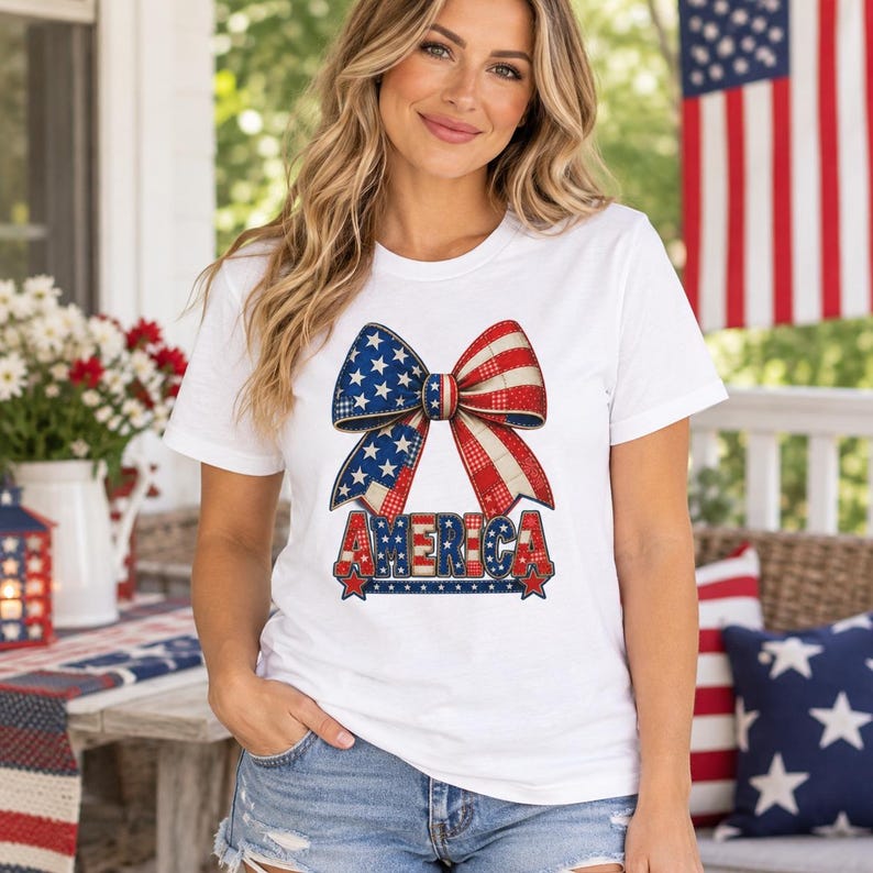 May include: A white short-sleeved t-shirt featuring a large graphic of a bow patterned with red, white, and blue stars and stripes. Below the bow, the word AMERICA is printed in bold, block letters with a matching patriotic star-spangled design.
