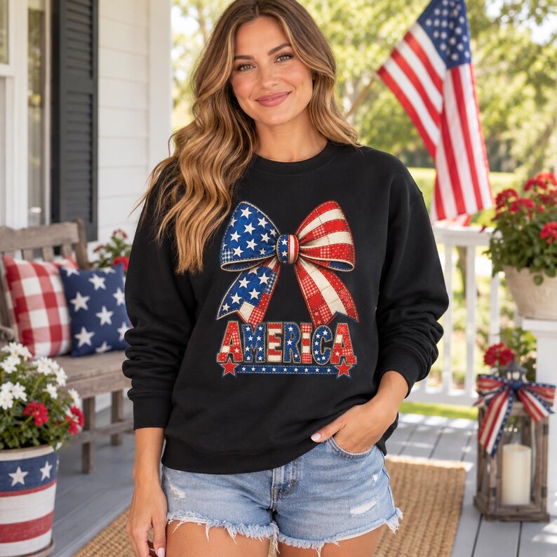 May include: A black long-sleeve sweatshirt featuring a large graphic of a bow with a red, white, and blue star and stripe pattern. Below the bow, the word AMERICA is printed in a matching patriotic pattern with small red stars at the ends.
