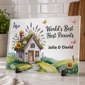 Pode incluir: Uma tábua de corte retangular de vidro com um design decorativo. O design apresenta uma pequena casa com telhado verde, girassóis e o texto "World's Best Host Parents" com os nomes Julia & David.