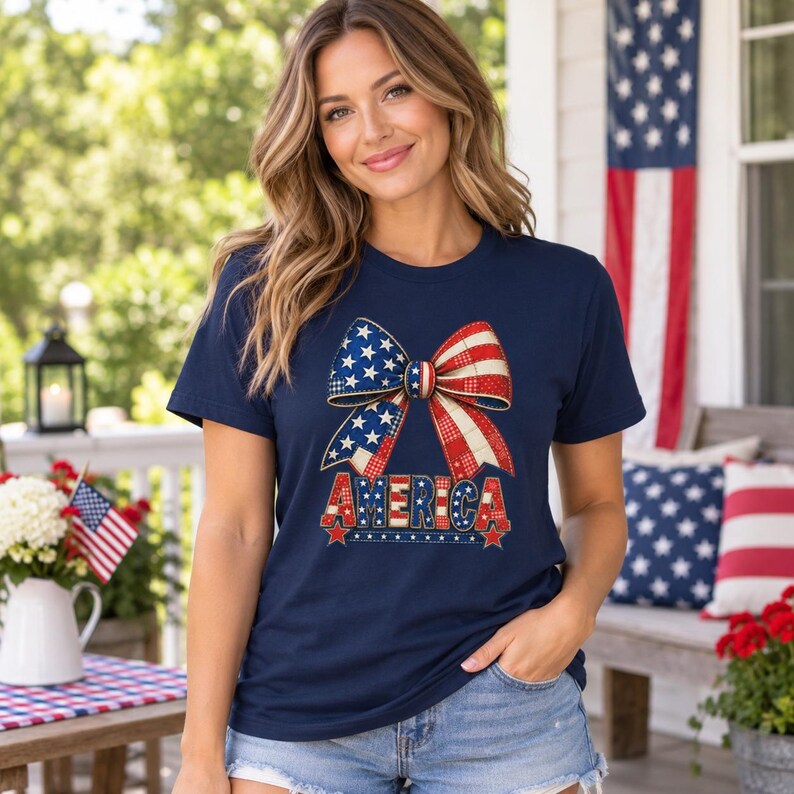 May include: A navy blue short-sleeve t-shirt featuring a large graphic of a bow patterned with red, white, and blue stars and stripes. Below the bow, the word AMERICA is printed in bold, block letters with a matching patriotic star and stripe design.