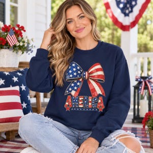 May include: A navy blue long-sleeve sweatshirt featuring a large graphic of a bow with a red, white, and blue star and stripe pattern. Below the bow, the word AMERICA is printed in a bold, block font with a matching patriotic color scheme.