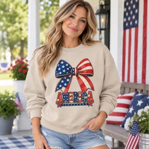 May include: A beige long-sleeve sweatshirt featuring a large graphic of a bow patterned with red, white, and blue stars and stripes. Below the bow, the word AMERICA is printed in bold, block letters with a matching patriotic color scheme.