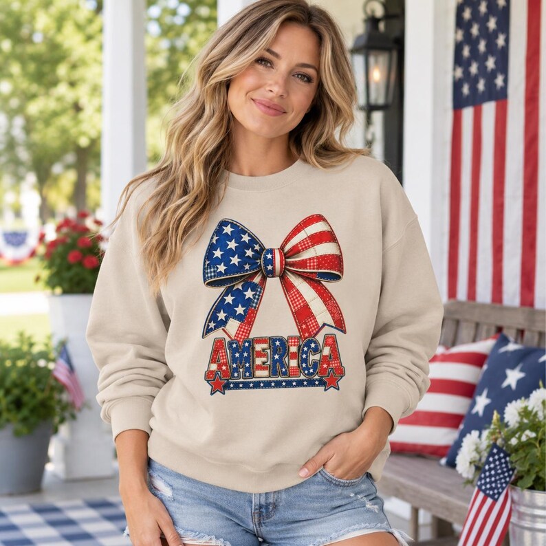 May include: A beige long-sleeve sweatshirt featuring a large graphic of a bow patterned with red, white, and blue stars and stripes. Below the bow, the word AMERICA is printed in bold, block letters with a matching patriotic color scheme.