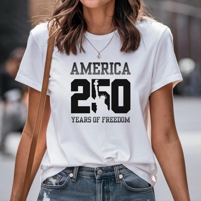Puede incluir: Camiseta blanca con el texto "AMERICA 250 YEARS OF FREEDOM" en negro y gris. El n&uacute;mero 250 est&aacute; en negrita, con la silueta de la Estatua de la Libertad incorporada en el cero.