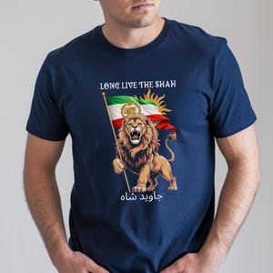 Può includere: T-shirt blu navy con un leone che tiene una bandiera. La bandiera ha strisce rosse, bianche e verdi, un sole e un emblema. È visibile anche il testo "LONG LIVE THE SHAH" e testo in farsi. Il leone è marrone chiaro con una criniera dettagliata.