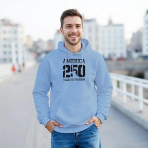 Puede incluir: Sudadera con capucha azul claro con el texto "AMERICA 250 YEARS OF FREEDOM" en negro. La sudadera tiene un bolsillo delantero y una capucha. La persona lleva vaqueros azules.