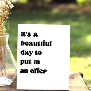 May include: White card with black text that reads "it's a beautiful day to put in an offer". The card is sitting on a wooden surface.