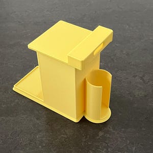 May include: A yellow 3D-printed desk organizer with a rectangular main body, a small tray, and a cylindrical holder. The organizer has a handle on top and is designed to hold various office supplies.