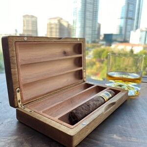 May include: A wooden cigar box with a hinged lid and three compartments. The box is open and a brown cigar is inside. The box is on a wooden surface.