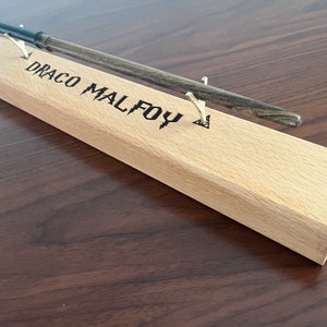 May include: A wooden wand display stand with the name "Draco Malfoy" engraved on the top. A black and gray wand is resting on the stand.