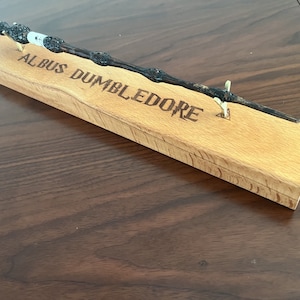 Könnte beinhalten: Ein schwarzer Zauberstab mit einem Holzständer. Der Ständer ist mit den Worten "Albus Dumbledore" graviert.