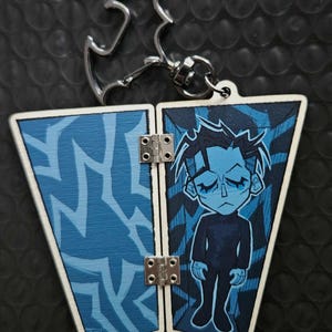 May include: A hinged, double-sided keyring featuring a cartoon figure. One side has a blue abstract design, the other a blue figure with black clothing. The keyring has a silver bat-shaped clasp.