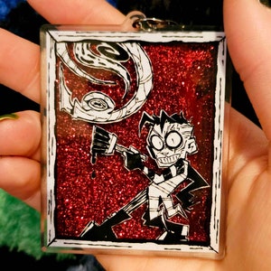 May include: A red glitter keychain featuring a black and white cartoon character holding a paintbrush. The character has a surprised expression and is wearing a striped shirt. The background is a red glitter with a black and white swirl design.