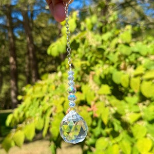 May include: A clear crystal ball suncatcher hanging from a silver chain. The crystal ball is faceted, reflecting light. Above the ball are iridescent beads. The background is blurred green foliage.