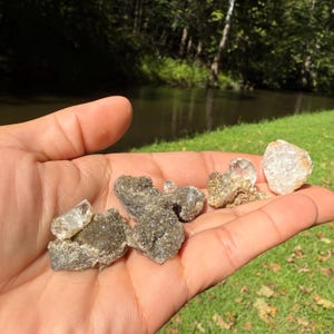 Puede incluir: Una colección de cuatro especímenes minerales en bruto y sin pulir que descansan en una palma abierta. Las piedras presentan puntas cristalinas translúcidas y claras que sobresalen de matrices rocosas rugosas, de color gris oscuro y textura terrosa.